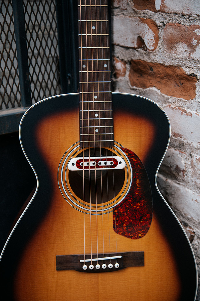 Close-up M-240E Antique Burst leaning in corner of brick wall and fence showing front body view with electronics
