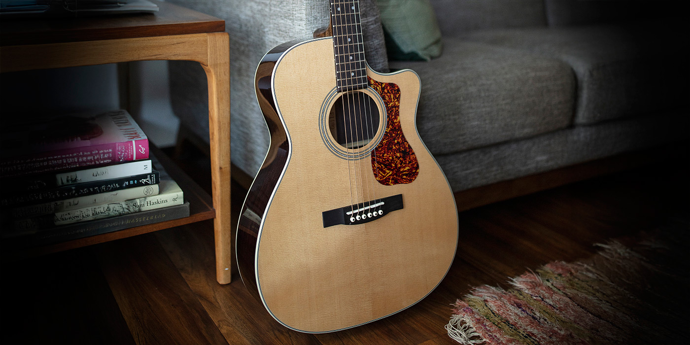 OM-150CE leaning against a cloth couch by an end table with a stack of books on bottom shelf