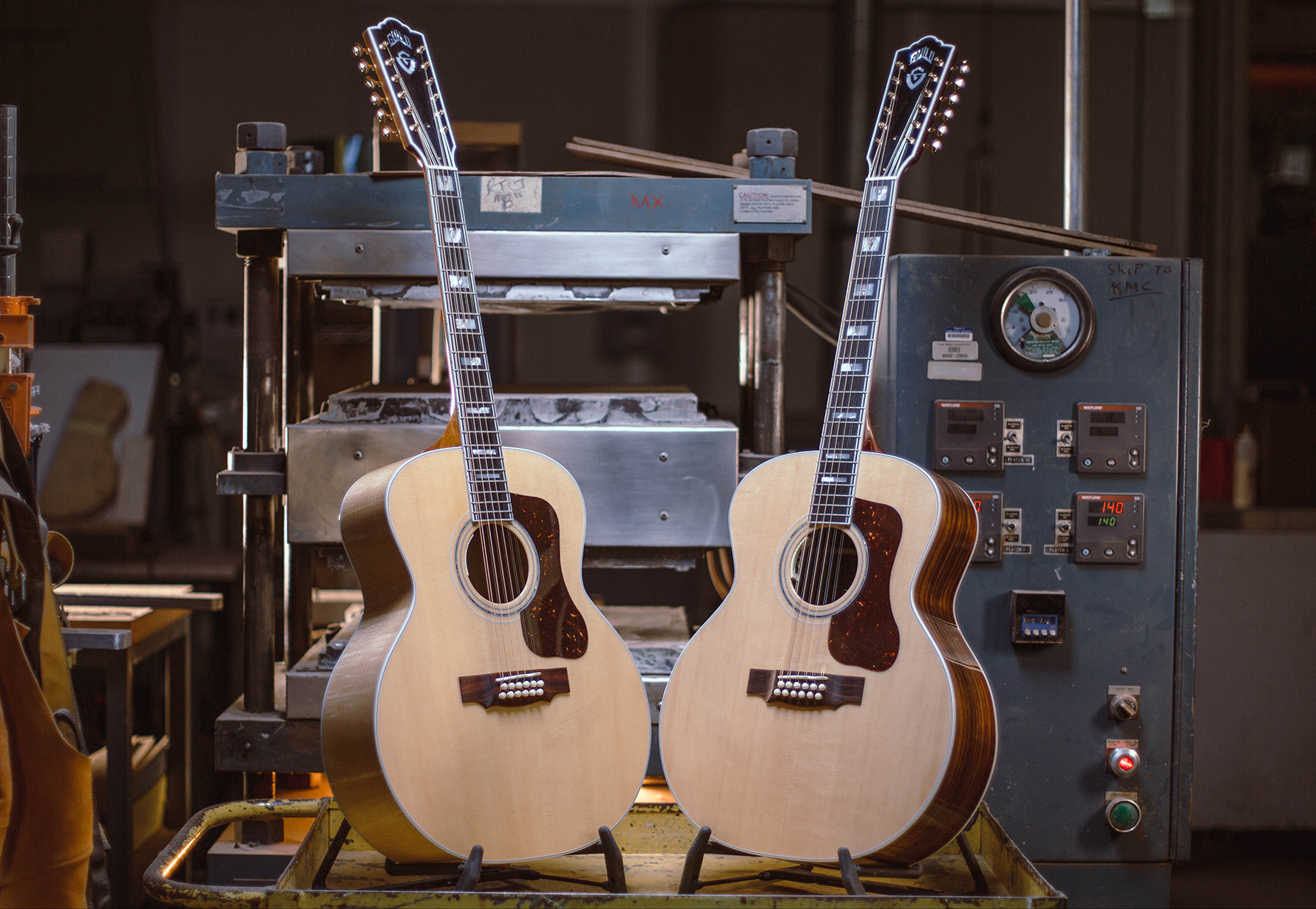 F-512 acoustic guitars are displayed in guitar workshop, with a machinery backdrop and tools nearby. The setting is well-lit and industrial.