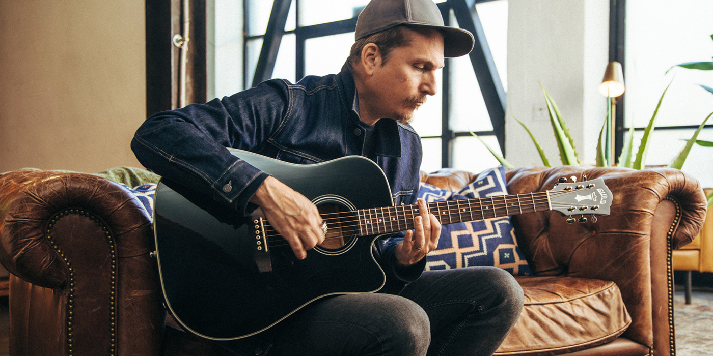 D-140CE Black being played by a guitarist sitting on a leather couch in an apartment
