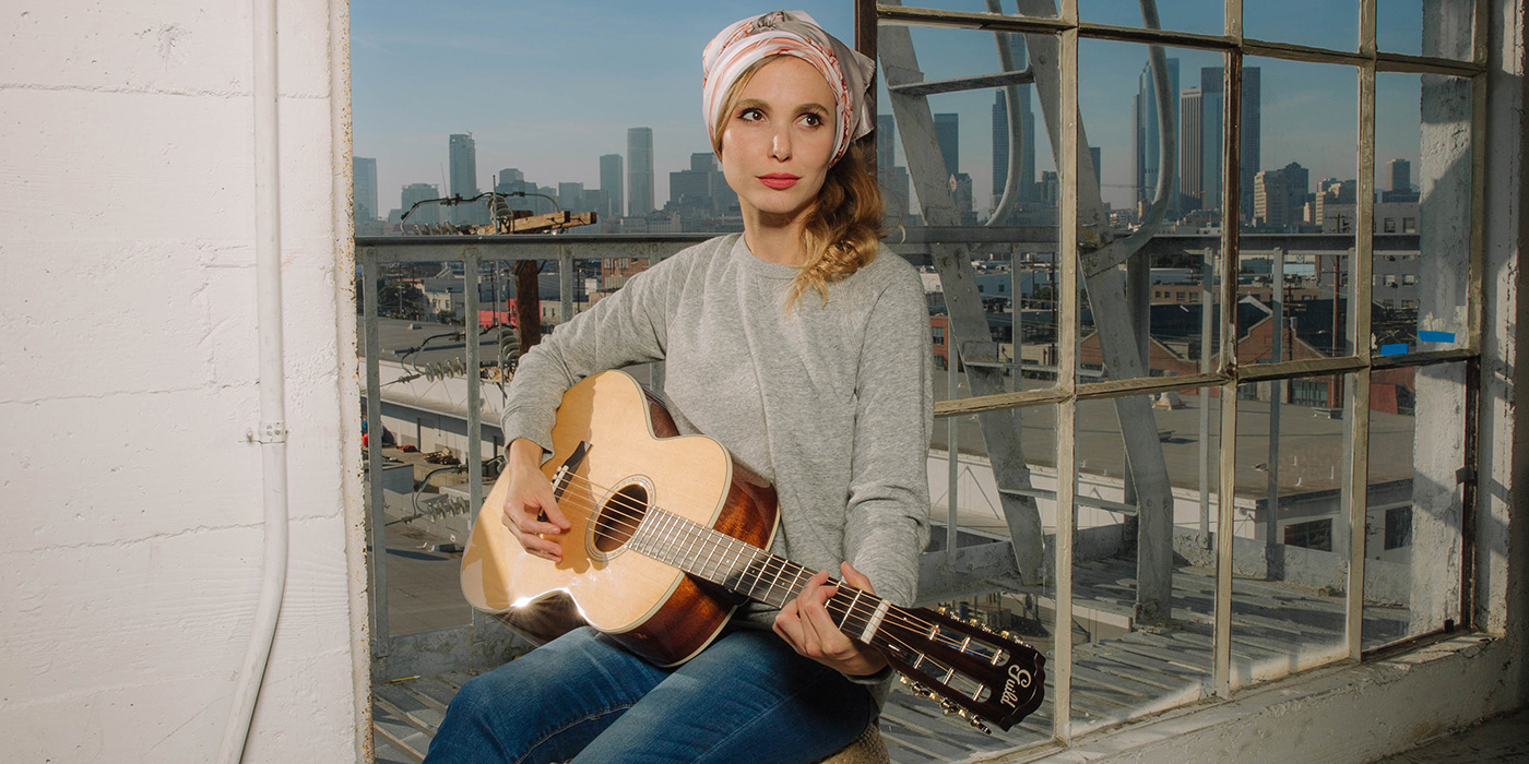 Woman playing a P-240 Memoir white sitting on the ledge of a window overlooking a city skyline.