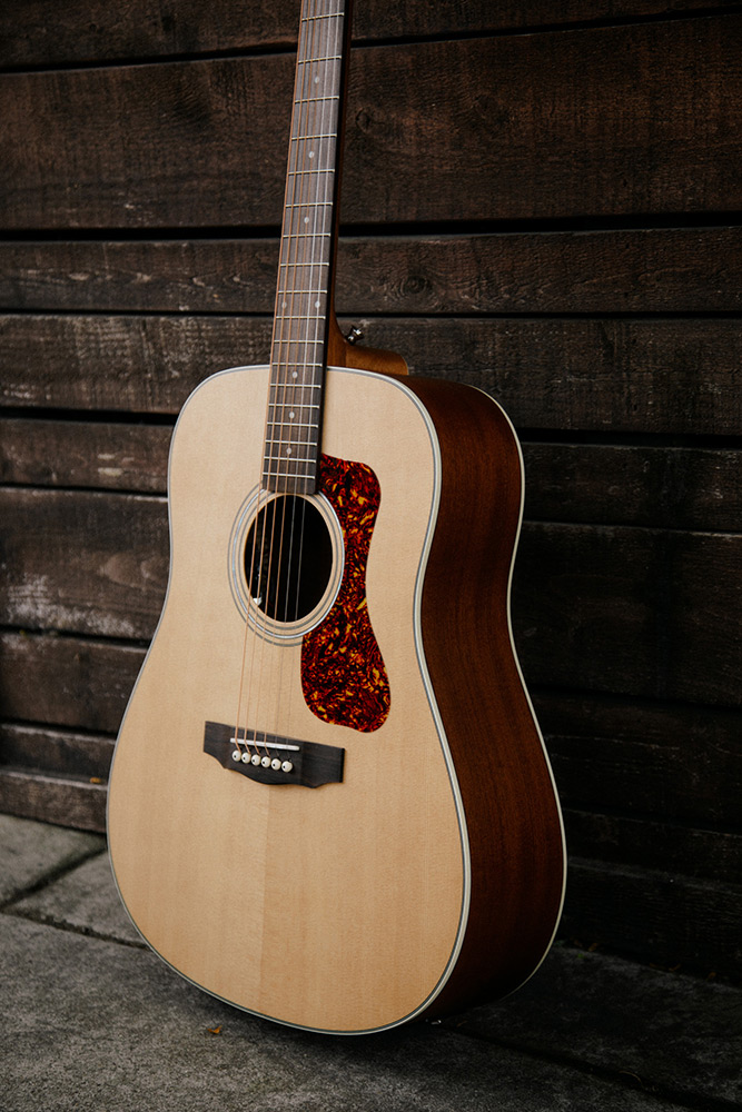 Close up D-240E Natural leaning against dark wood fence showing pickguard, soundhole, bridge, and Solid Sitka Spruce top