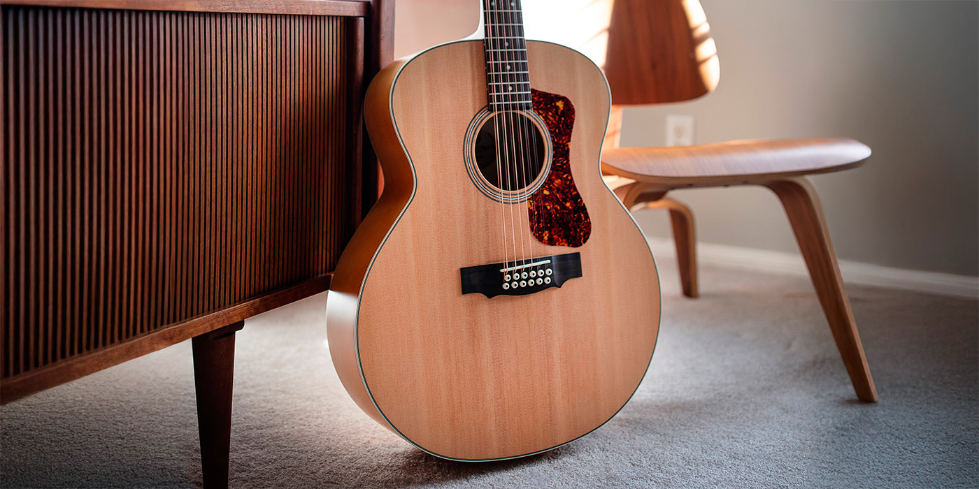 F-2512E Maple acoustic guitar in front of a modern wood cabinet and chair