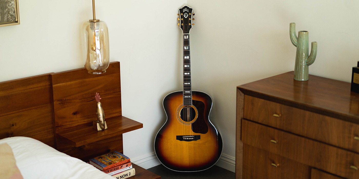 F-55E Maple Antique Burst acoustic guitar leans against a wall in a cozy room, accompanied by a bedside lamp and decorative items on furniture.