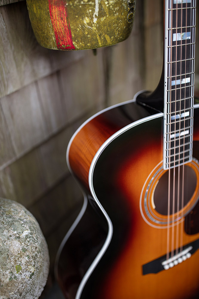A close-up of F-55E Maple acoustic guitar with a warm, antique burst finish, leaning against a textured wall and near a stone.